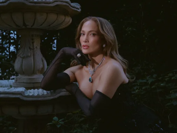 Η Jennifer Lopez ποζάρει με κοστούμι και κοσμήματα δίπλα σε ένα σιντριβάνι, με φόντο πλούσια βλάστηση.