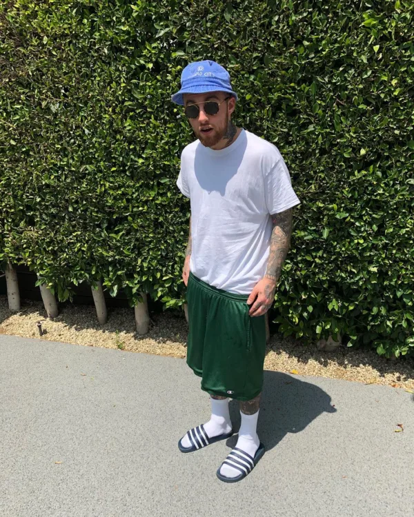 Ο Mac Miller με μπλε καπέλο και πράσινες βερμούδες στέκεται μπροστά από φράχτη, με αφορμή την επέτειο του «The Divine Feminine».