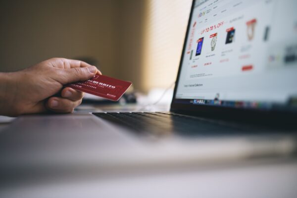 Χέρι κρατά πιστωτική κάρτα μπροστά από οθόνη laptop με ιστοσελίδα online αγορών, δείχνοντας πώς το κινητό χειραγωγεί την πιστωτική κάρτα.