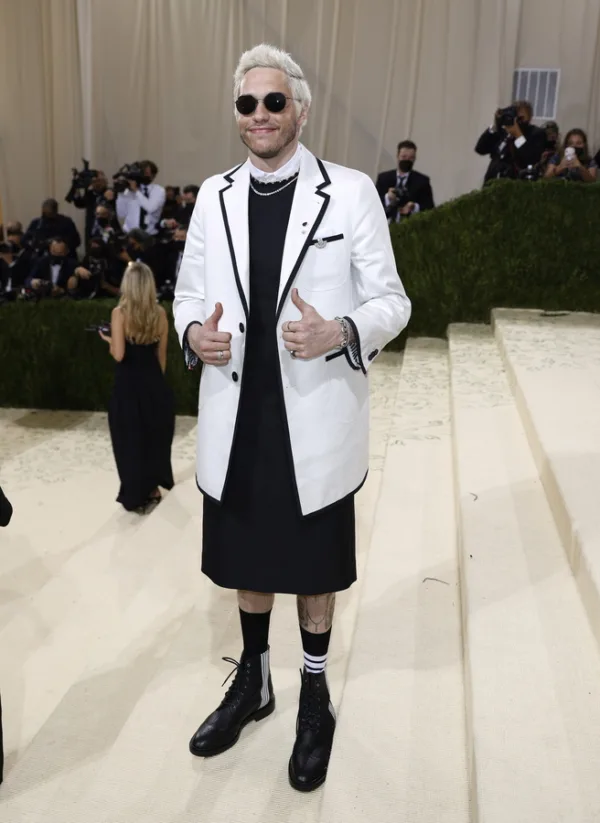 Ο Pete Davidson δίνει το σήμα της νίκης με τα δύο του δάχτυλα, φορώντας ένα λευκό σακάκι και μαύρο φόρεμα, ενώ δέχεται σχόλια.