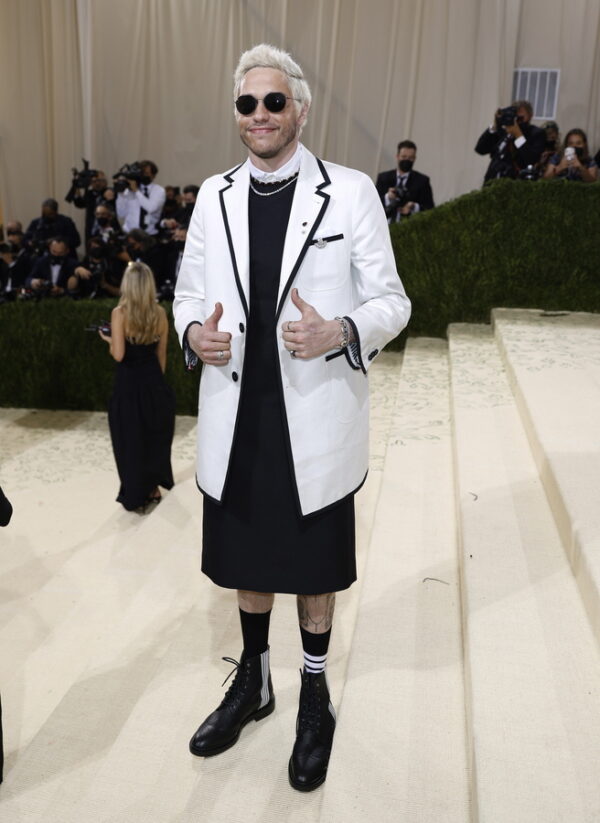 Ο Pete Davidson δίνει το σήμα της νίκης με τα δύο του δάχτυλα, φορώντας ένα λευκό σακάκι και μαύρο φόρεμα, ενώ δέχεται σχόλια.